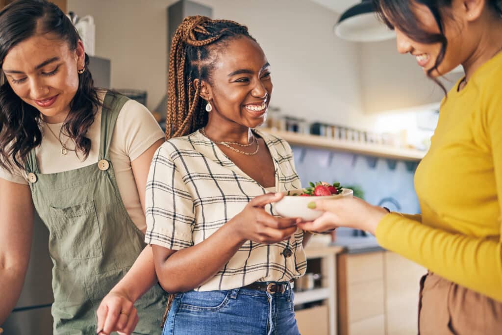 Three women in a kitchen, one handing a bowl of strawberries to another